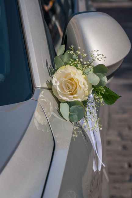 A close-up of a white rose with lush green eucalyptus leaves and delicate sprigs of baby's breath, attached to the side mirror of a silver car. The bouquet features fresh, soft petals and slender stem