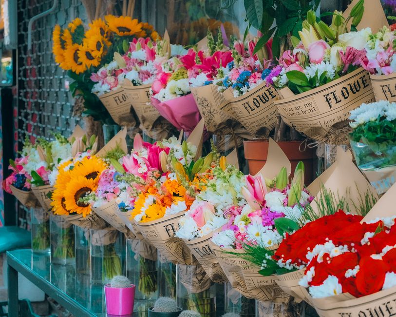 An array of fresh floral bouquets displayed on a glass shelf at King Street florists Twickenham. Each bouquet is wrapped in brown, newspaper-style paper with floral printing and tied with twine or rib
