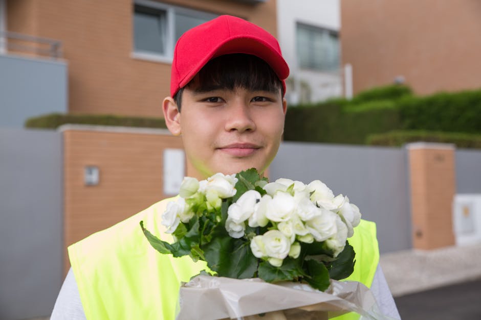 A young male flower delivery courier wearing a red cap and high-visibility yellow vest is holding a bouquet of white, fully bloomed flowers, likely roses or similar blossoms, wrapped in white paper. H
