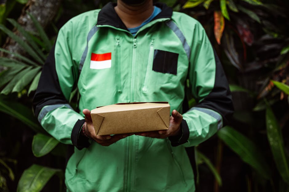 A delivery person, wearing a green jacket with reflective strips and a flag patch, holds a small, plain brown paper package with folded corners in both hands. The individual is standing in front of lu